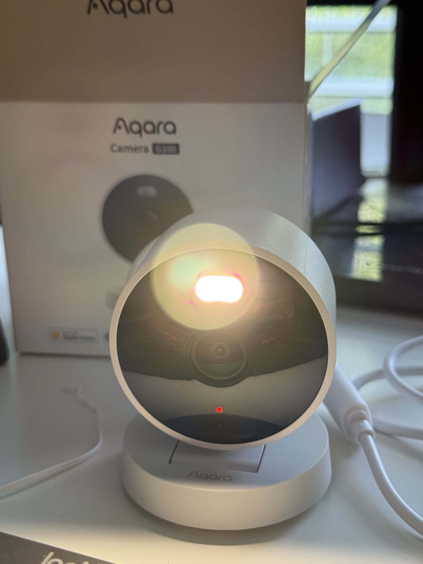 The Aqara G100 powered on in front of its box, with its front spotlight glowing and a small red status LED lit beside the lens.