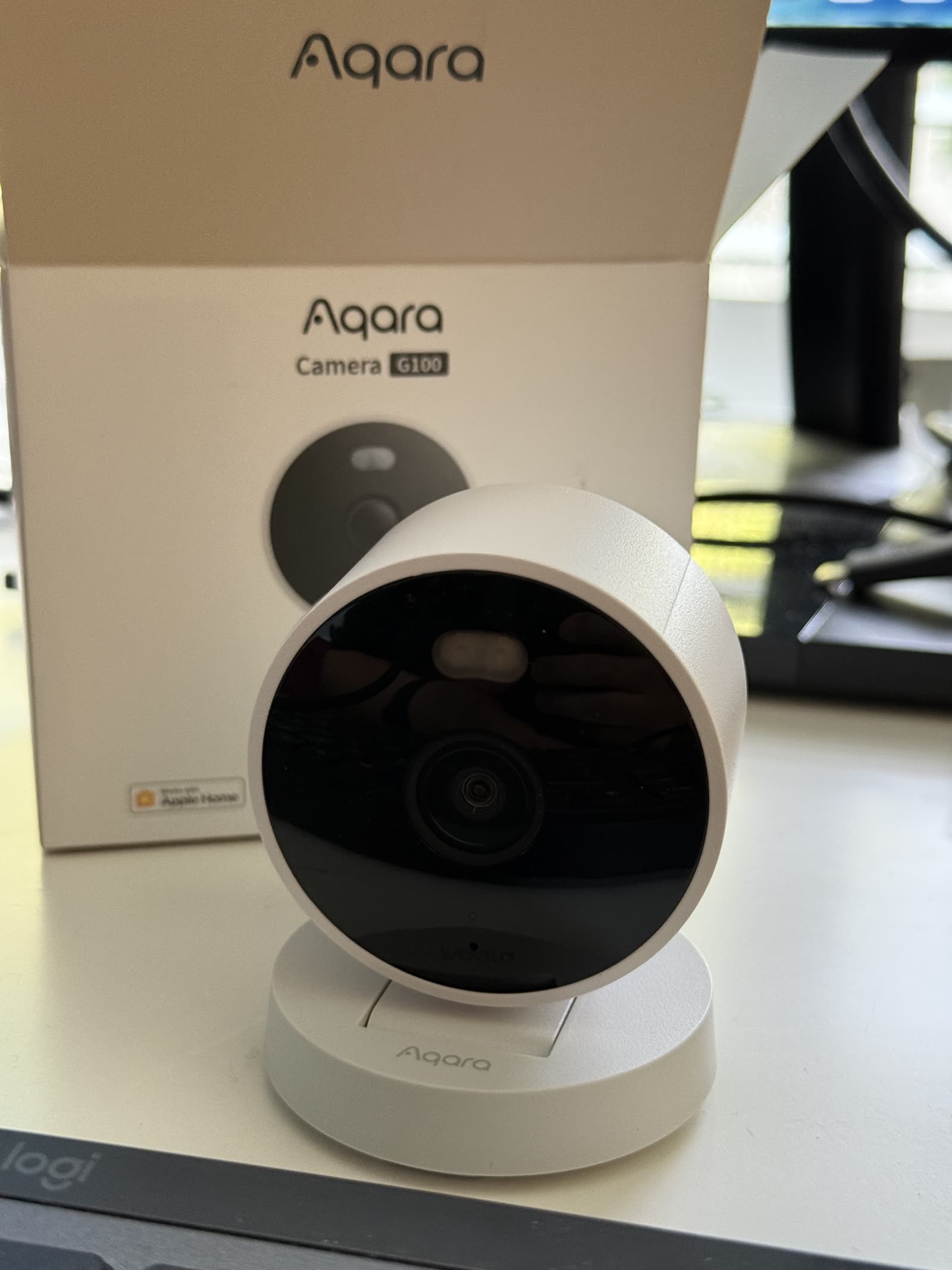 The white Aqara Camera G100 on its round stand in front of its box, which carries the Aqara logo, the product name, and a Works with Apple Home badge.