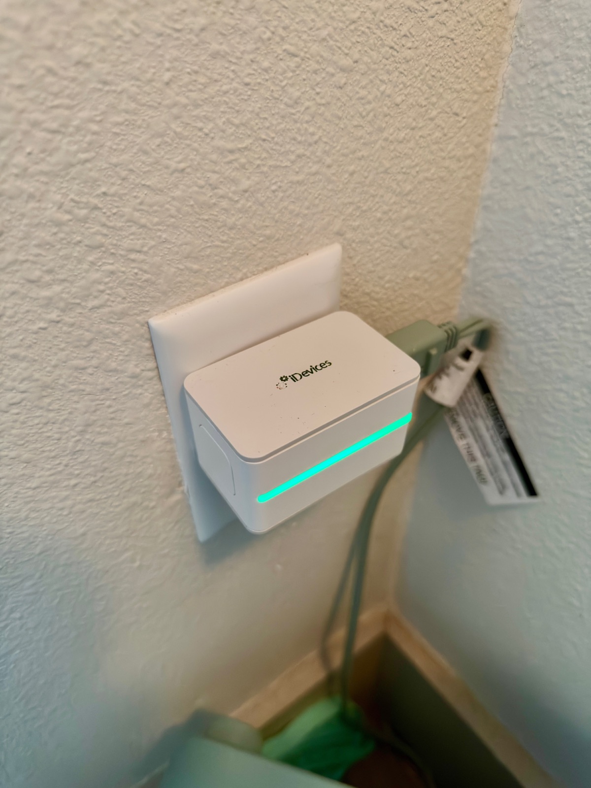 An iDevices Switch smart plug in a wall outlet with its green LED night light strip lit along the bottom edge, and a cable plugged into the side-facing socket on the right so the cord runs flat down the wall instead of jutting out.