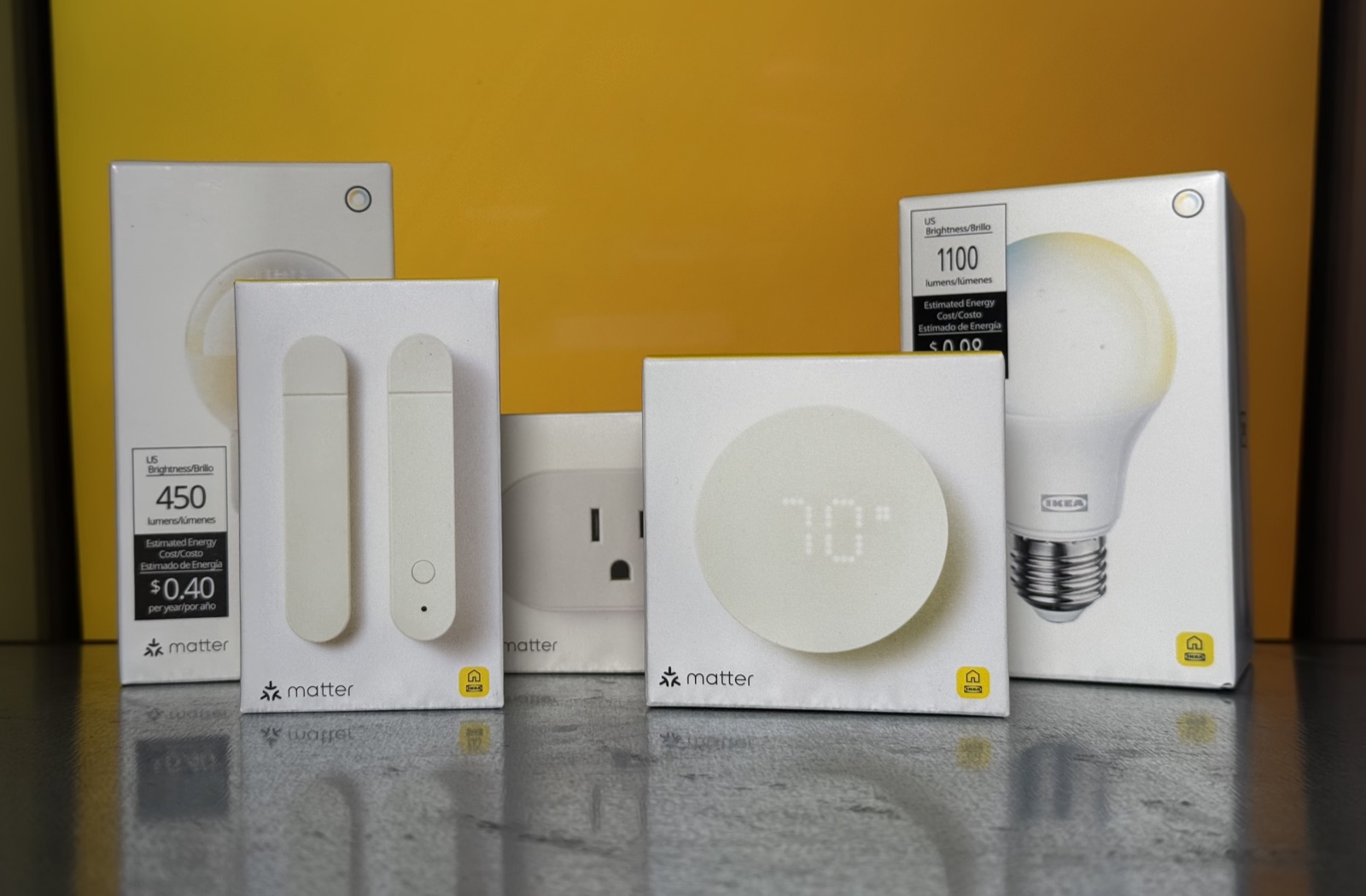 Five IKEA smart home boxes on a counter against a yellow background, each carrying the Matter logo: a 450 lumen bulb, a two-piece sensor, a smart plug showing a US outlet, a round display unit reading 70 degrees, and an 1100 lumen A19 bulb.