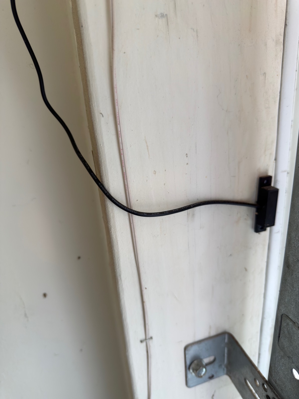 The sensor cable running down the painted door jamb to the black reed switch mounted on the frame, held flat against the trim.