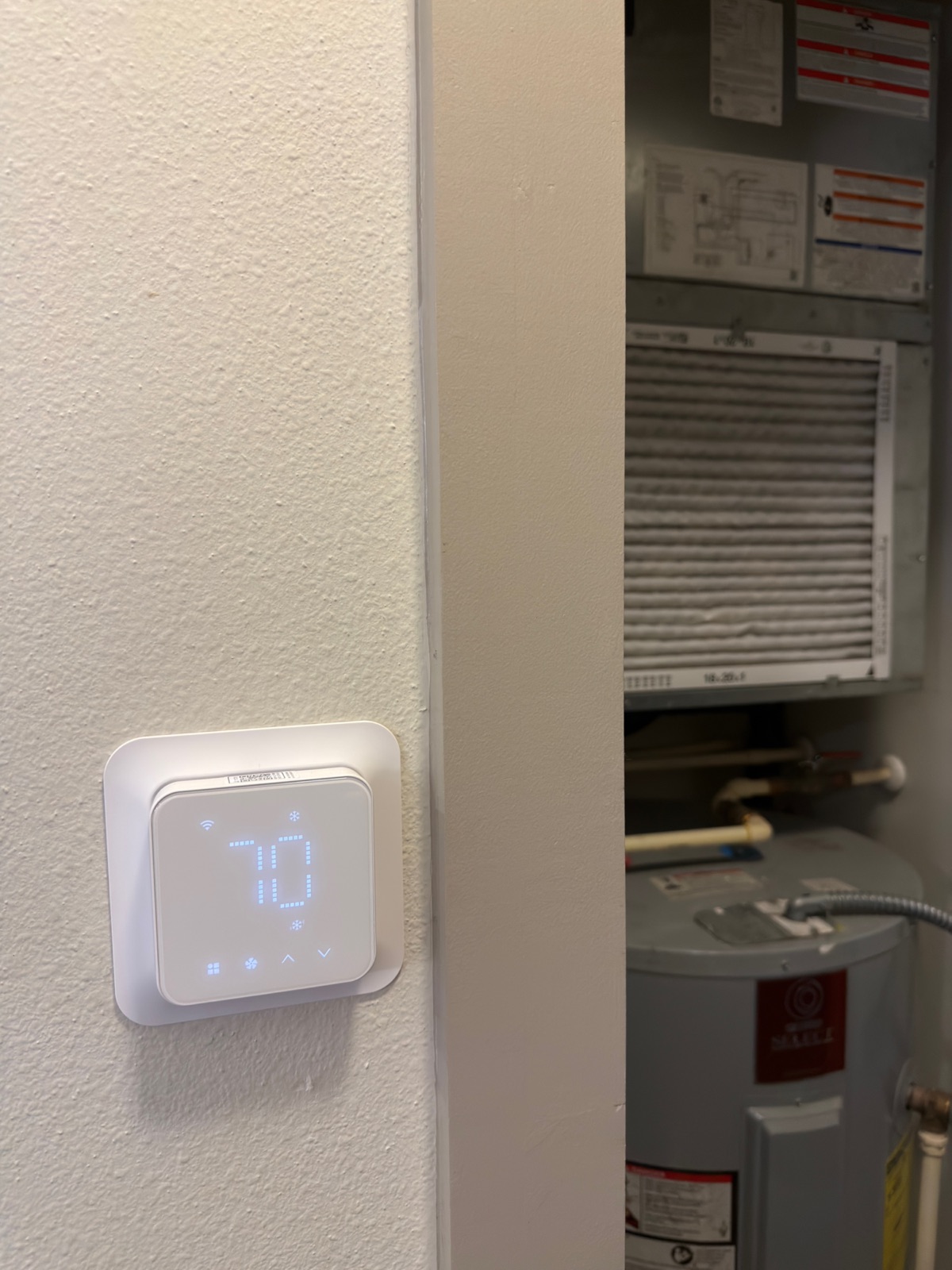The Meross Matter thermostat mounted on a hallway wall showing 70 degrees, with the open air handler closet beside it: the return filter, the equipment wiring label, and the water heater below.