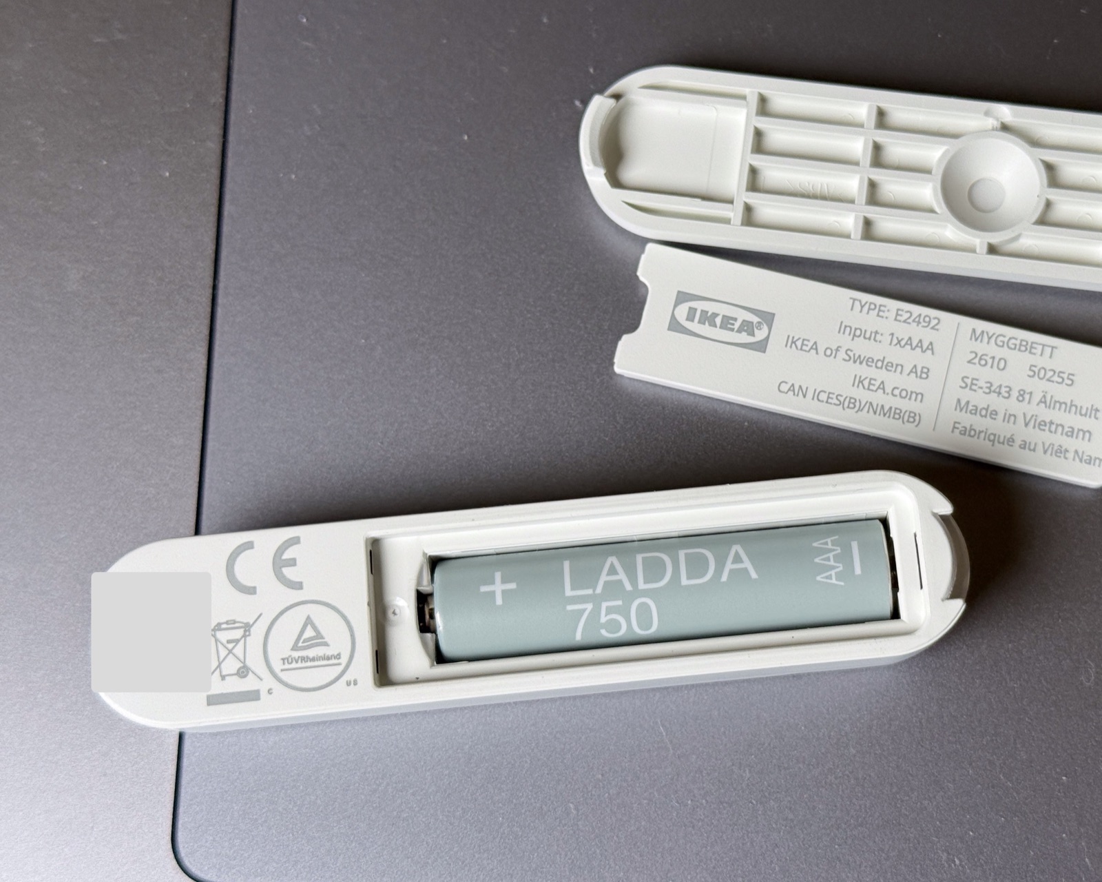 The MYGGBETT opened for battery installation, showing a LADDA 750 AAA cell in the compartment and the labeled inner door, with the QR code area obscured.