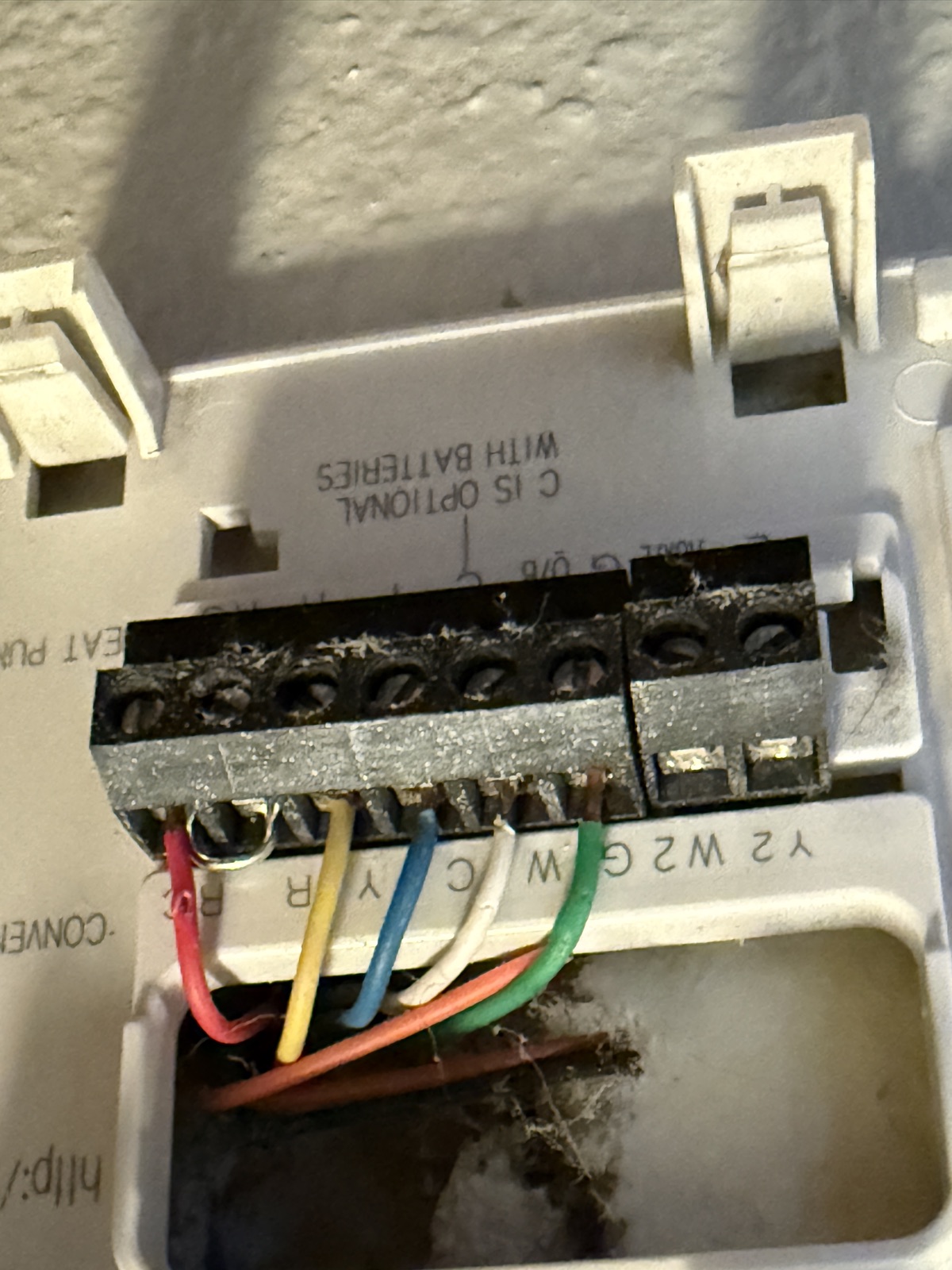 The old Honeywell thermostat back plate with wires still attached. Terminals are labelled Rc, R, Y, C, W and G down the strip, with a red wire on Rc, yellow on Y, blue on C, white on W and green on G. Printed text on the plate reads C IS OPTIONAL WITH BATTERIES.